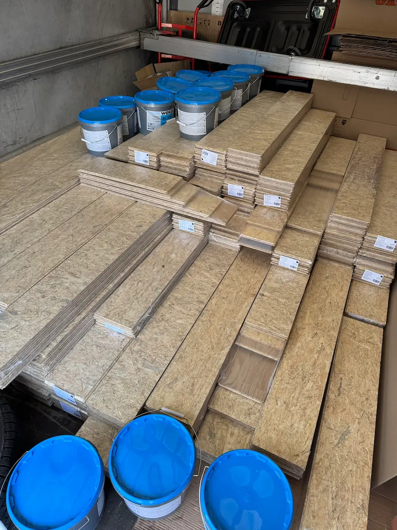 Stosy drewnianych płyt i niebieskie wiadra z klejem montażowym na pace ciężarówki
